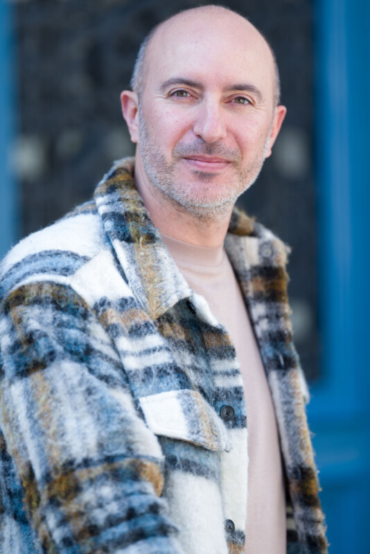 A middle-aged man with a close-shaved head and beard, wearing a beige shirt and a plaid, fuzzy jacket, stands outdoors in front of a blurred, blue and dark background, looking confidently at the camera.