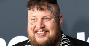A smiling man with a short curly haircut, beard, and multiple face tattoos, wearing a black and white patterned shirt, stands in front of a dark background.