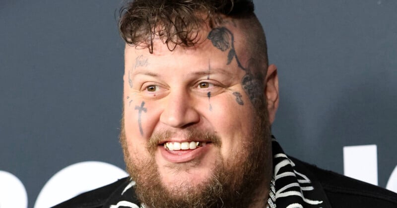 A smiling man with a short curly haircut, beard, and multiple face tattoos, wearing a black and white patterned shirt, stands in front of a dark background.