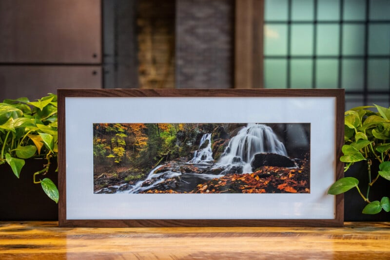 Una foto de paisaje enmarcada de una cascada rodeada de hojas de otoño sentadas en una mesa de madera flanqueada por plantas verdes en macetas con un fondo interior suave moderno.