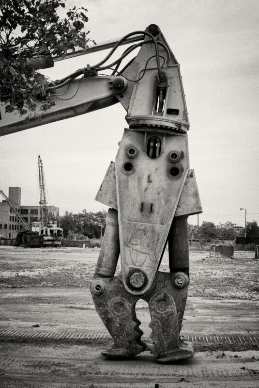 A large excavator attachment with a jaw-like structure stands upright at a construction site, resembling a robot; cranes and buildings are visible in the background under a cloudy sky.