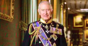 An older man with gray hair stands indoors wearing a formal, ornate military uniform adorned with medals, ribbons, and sashes, with a richly decorated background and framed artwork.