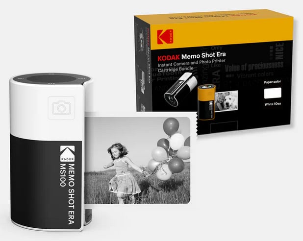 A Kodak Memo Shot Era MS100 instant camera and photo printer prints a black-and-white photo of a girl with balloons; the product box is in the background showing branding and cartridge information.