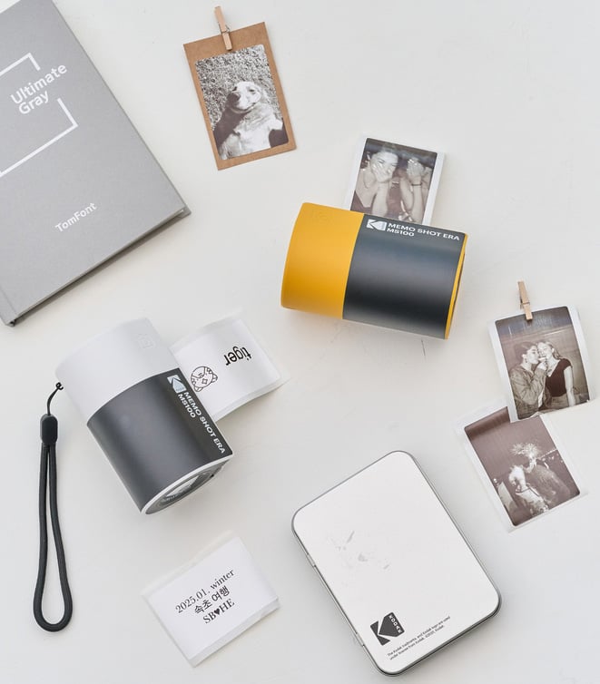 A flat lay of instant photos, a gray notebook labeled "Ultimate Gray," two compact Kodak instant photo printers, a white card, and a small white case on a light surface.