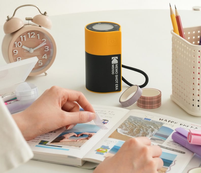 A person decorates a journal with photos and tape at a desk. Nearby are an alarm clock, a small printer, washi tapes, and a white basket holding pens. The scene looks bright and organized.