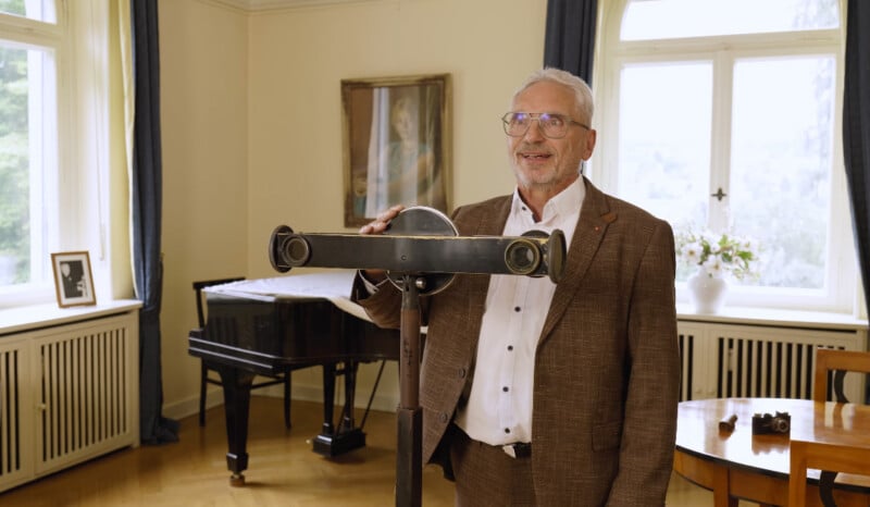 An older man in a brown suit stands indoors, smiling beside a vintage optical device on a stand. Behind him are large windows, a grand piano, and framed photos on the window ledge.