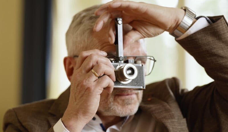 An older man with gray hair and glasses is holding a vintage camera up to his eye, preparing to take a photograph. He is wearing a brown jacket and a gold ring.