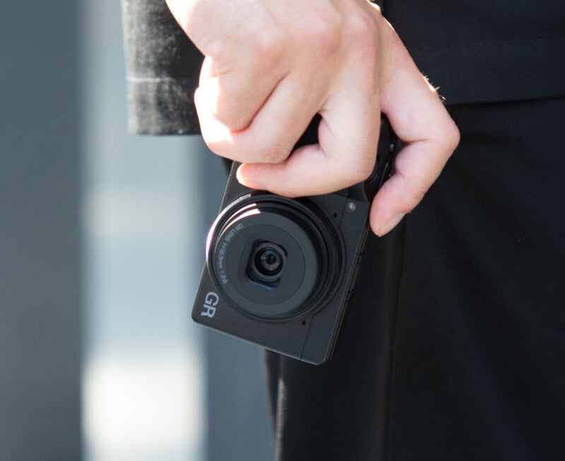 A person holds a compact Ricoh GR digital camera in their right hand, with the lens facing forward and their black clothing visible in the background.