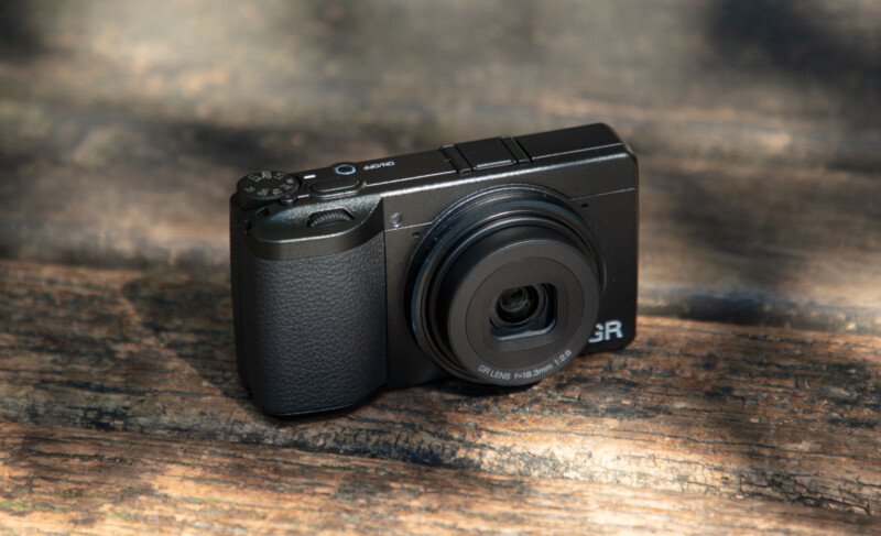 A compact black digital camera with a protruding lens rests on a weathered wooden surface, illuminated by natural sunlight.