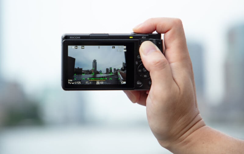 A hand holds a digital camera, focusing on a cityscape with tall buildings, as seen on the camera screen; the background is blurred and out of focus.