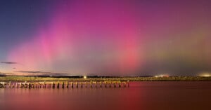A vivid aurora with pink and purple lights illuminates the night sky above calm water and a wooden pier, with a rocky breakwater in the background.