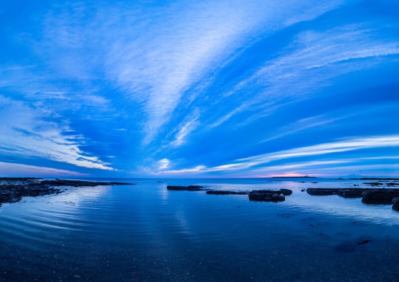 A wide view of a calm sea at sunset with dramatic blue clouds streaking across the sky, reflecting on the water. Some rocks are visible near the shore. The horizon glows with hints of orange and pink.