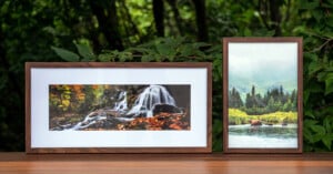 Two framed nature photographs on a wooden surface outdoors; one shows a waterfall with autumn leaves, the other shows a bear walking by a lake with pine trees and misty mountains in the background.