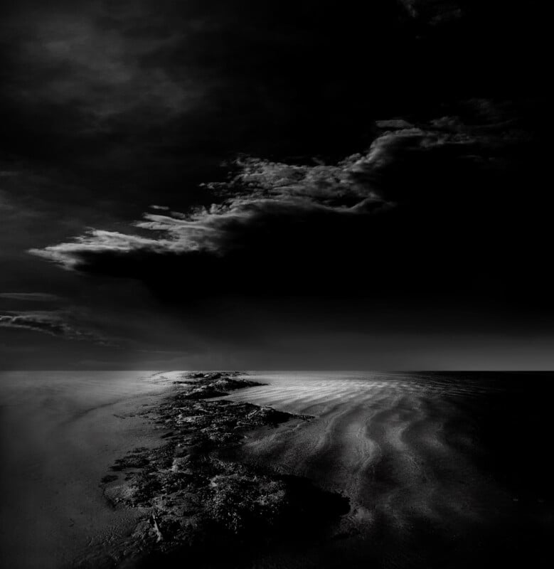 A dramatic black and white seascape shows a rocky shoreline stretching into the distance, with dark, textured clouds looming above a rippled, sandy beach under moody lighting.
