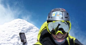A climber in bright gear and reflective goggles smiles for a selfie near a snowy mountain peak under a clear blue sky. Snow is kicked up in the air, and equipment is visible behind the climber.