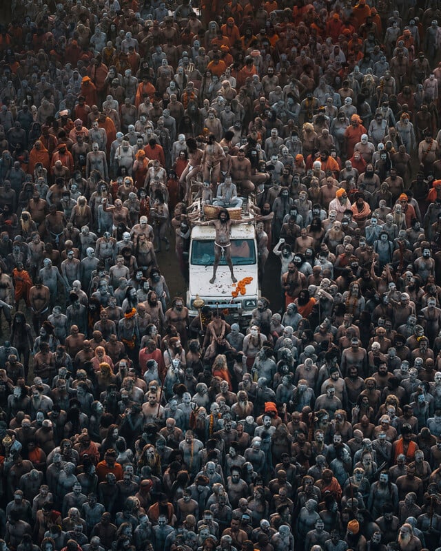 A large crowd of people, many with their bodies covered in ash, surrounds a white vehicle topped with individuals, forming a dense gathering at what appears to be a religious or cultural event.