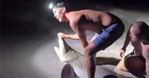 Two men, one wearing a headlamp, crouch on the sand at night, holding a large shark near the water's edge. The man in front is smiling and gripping the shark's head while the other supports from behind.