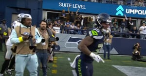 A Seattle Seahawks player runs along the sideline during a football game as photographers and fans watch from behind the barrier. The background shows excited fans and a "Suite Level" sign.