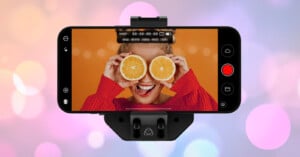 A smartphone on a mount displays a woman holding two orange halves over her eyes and smiling, with an orange background, while the phone screen shows camera controls.