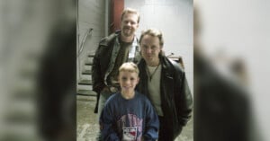 Three people pose together indoors near a staircase. Two adults in leather jackets stand behind a smiling young boy wearing a blue New York sweatshirt. The background features concrete walls and metal stairs.