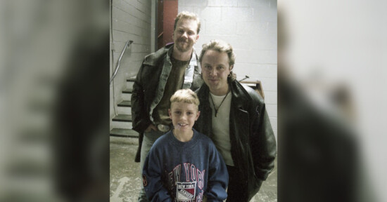 Three people pose together indoors near a staircase. Two adults in leather jackets stand behind a smiling young boy wearing a blue New York sweatshirt. The background features concrete walls and metal stairs.