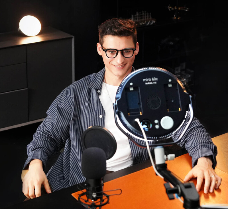 A smiling person wearing glasses sits at a desk with a microphone and ring light, recording or streaming video content in a modern, dimly lit room.