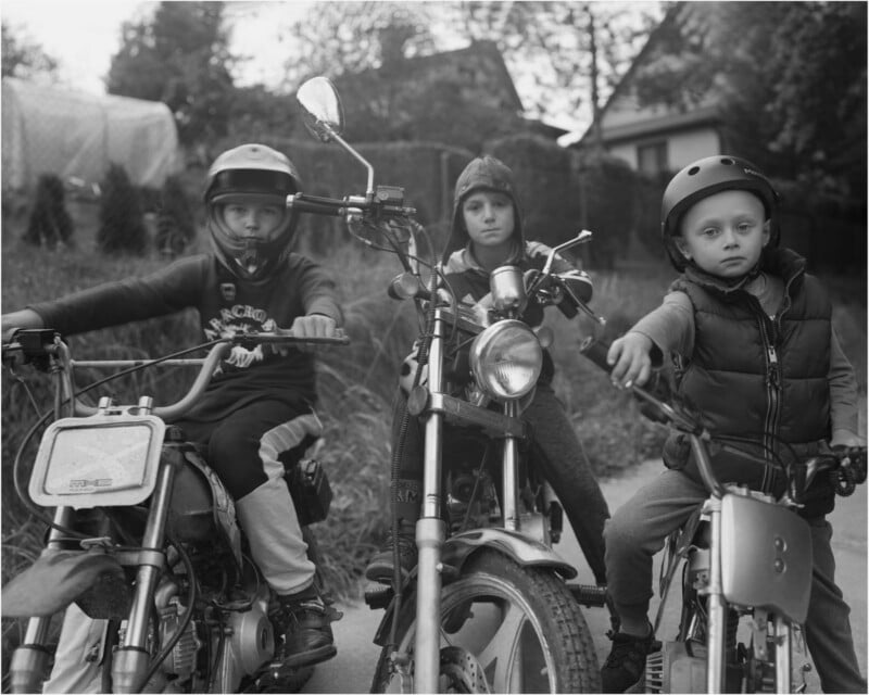 Three children wearing helmets sit on motorbikes and mopeds on a path with houses and trees in the background. They look directly at the camera with serious expressions. The image is in black and white.