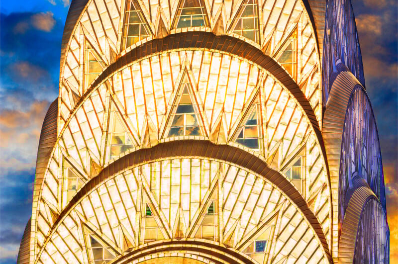 Close-up view of the illuminated Art Deco crown and spire of the Chrysler Building in New York City, with geometric patterns glowing against a dramatic evening sky.