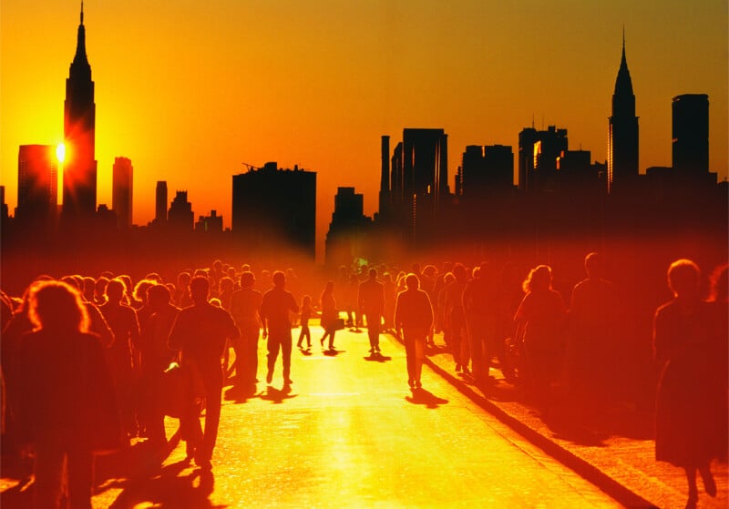 Crowds of people walk along a sunlit street with the New York City skyline, including the Empire State Building, silhouetted against a vivid orange sunset.