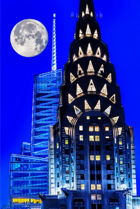 The illuminated Chrysler Building and a modern glass skyscraper stand against a vivid blue night sky, with a bright full moon visible in the upper left corner.