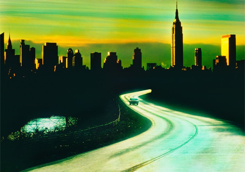 A single car drives on a curving road toward a city skyline at sunset, with tall buildings silhouetted against a yellow-green sky and reflective water visible on the left side of the road.
