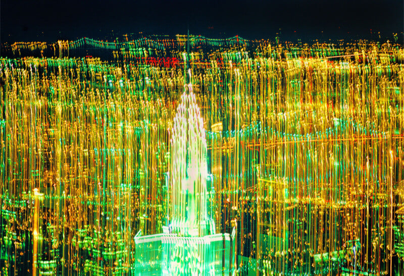 A nighttime cityscape with bright, colorful lights streaked vertically, creating an abstract effect. A tall, illuminated building is centered, surrounded by blurred yellow and green lines of city lights.