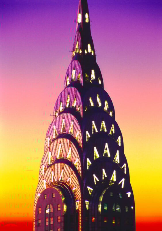 The top of the Chrysler Building in New York City is shown against a vivid gradient sunset sky in shades of yellow, orange, purple, and pink. The building’s illuminated Art Deco spire is prominently featured.