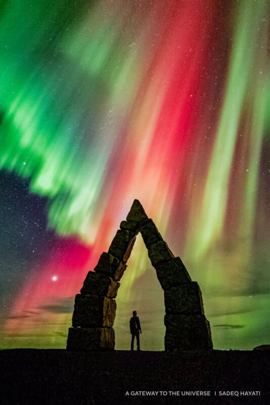 A person stands beneath a stone archway at night, silhouetted against vibrant red, green, and yellow aurora borealis lights swirling across a starry sky.