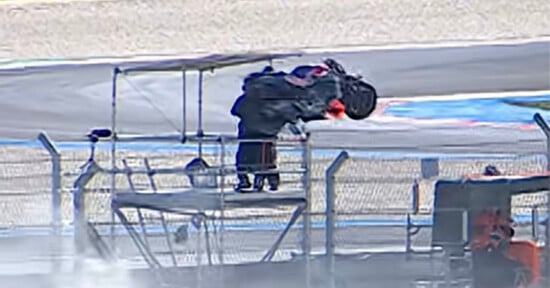 A race car is airborne after crashing, flipping over a safety barrier on a racetrack, with track officials nearby reacting to the accident.