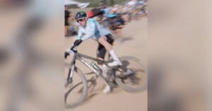 A cyclist wearing a helmet and sunglasses rides a mountain bike quickly on a dirt trail, with the motion creating a blurred effect. Spectators and tents are visible in the background.