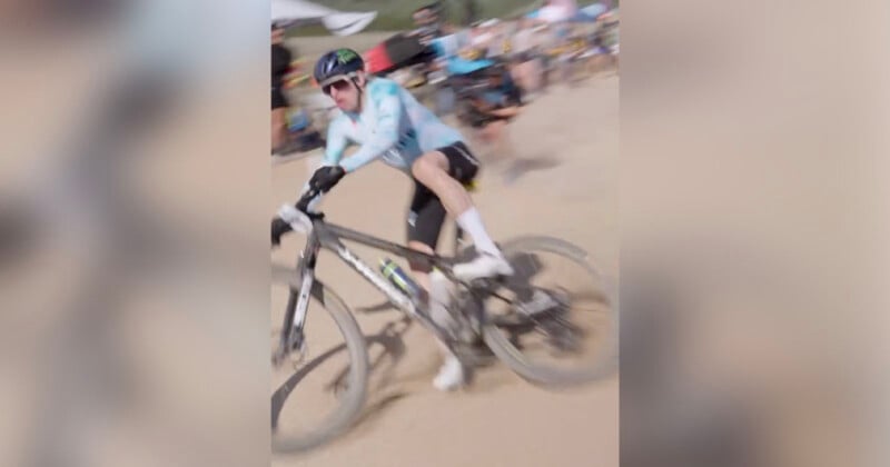 A cyclist wearing a helmet and sunglasses rides a mountain bike quickly on a dirt trail, with the motion creating a blurred effect. Spectators and tents are visible in the background.