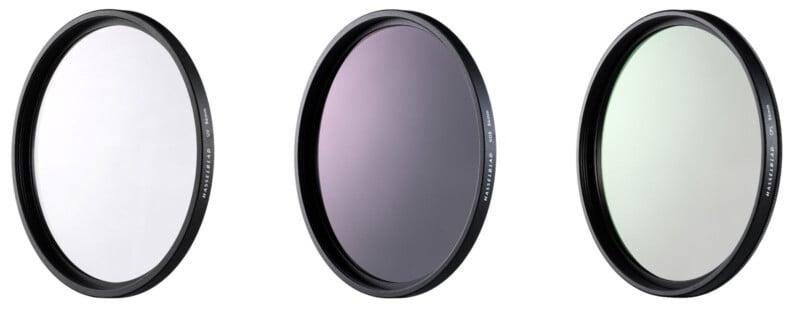 Three circular camera lens filters with black rims are displayed side by side on a white background; each filter has a slightly different tint—clear, purple, and greenish.