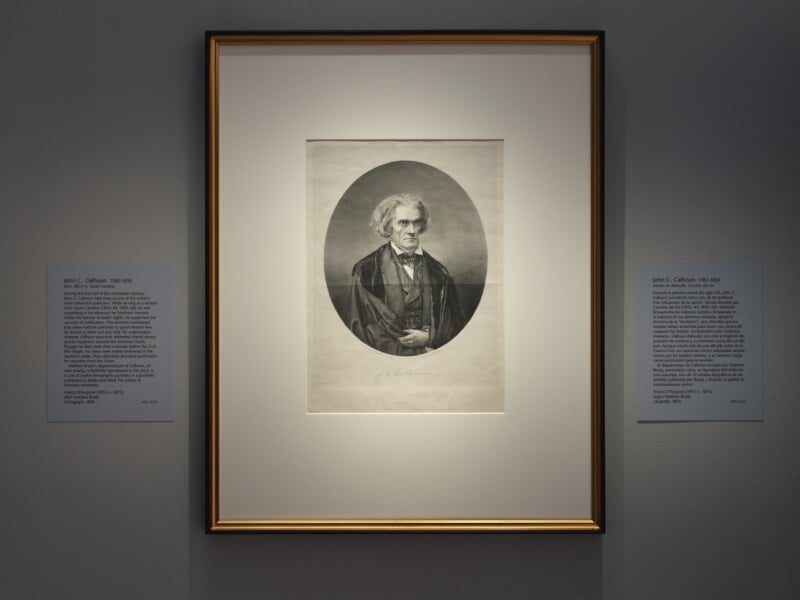 A framed black-and-white portrait of an older man in formal attire, displayed on a gallery wall with two informational plaques on either side of the frame.