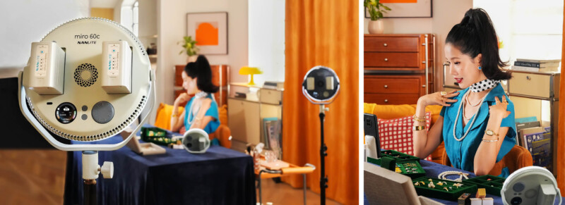 A woman wearing blue and orange clothing sits at a table displaying jewelry in a well-lit room, using ring lights and a camera to livestream or record herself showcasing various jewelry pieces.