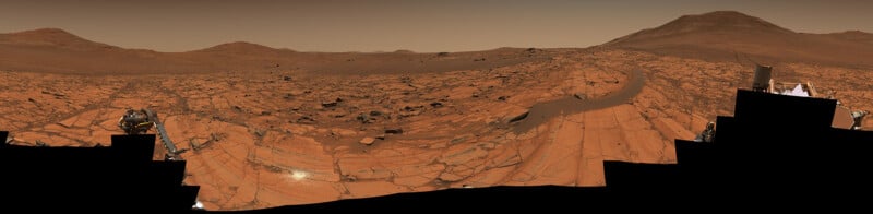 A panoramic view of the rocky, reddish Martian landscape with scattered hills and mountains under a hazy sky, captured by a rover with visible rover parts at the bottom edges of the image.