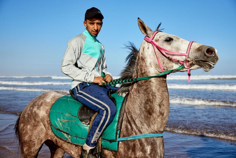 A young man wearing a cap and sports clothes rides a gray horse with a turquoise saddle and pink bridle along a beach, with waves and blue sky in the background.