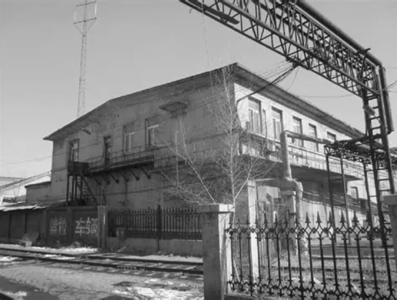 A black-and-white photo of an old, two-story industrial building with metal pipes, a fire escape, and a wrought-iron fence in front. Snow patches and bare trees are visible along a railway track.