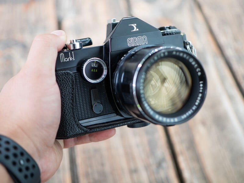 A hand holds a vintage Sigma Mark I film camera with a large lens, shown against a blurred wooden background.
