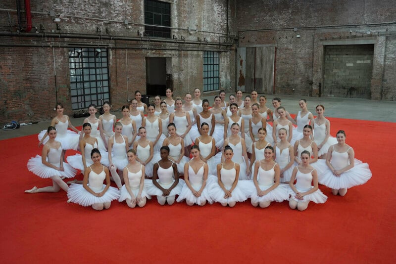 A large group of ballet dancers poses together on a red floor in an industrial-style studio, wearing white leotards and tutus. They are arranged in neat rows, smiling at the camera.