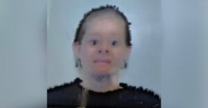 A blurry image of a person with short hair looking directly at the camera, wearing a dark top, against a plain light background. The facial features are indistinct due to the low resolution or distortion.