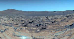Rocky landscape with cracked, layered ground and scattered boulders under a clear blue sky. Distant hills and mountains are visible on the horizon, creating a barren, desert-like scene.