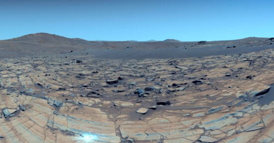 Rocky landscape with cracked, layered ground and scattered boulders under a clear blue sky. Distant hills and mountains are visible on the horizon, creating a barren, desert-like scene.