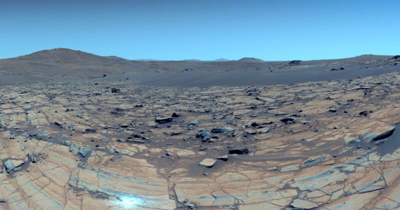 Rocky landscape with cracked, layered ground and scattered boulders under a clear blue sky. Distant hills and mountains are visible on the horizon, creating a barren, desert-like scene.
