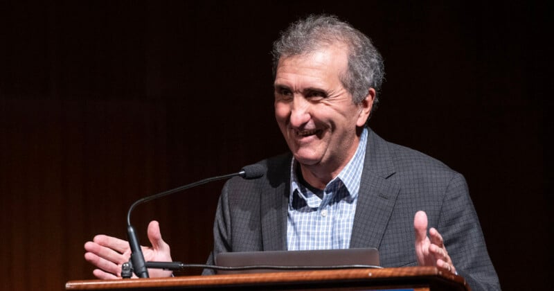 A man with gray hair wearing a checked shirt and dark blazer stands at a podium, smiling and gesturing with his hands while speaking into a microphone.